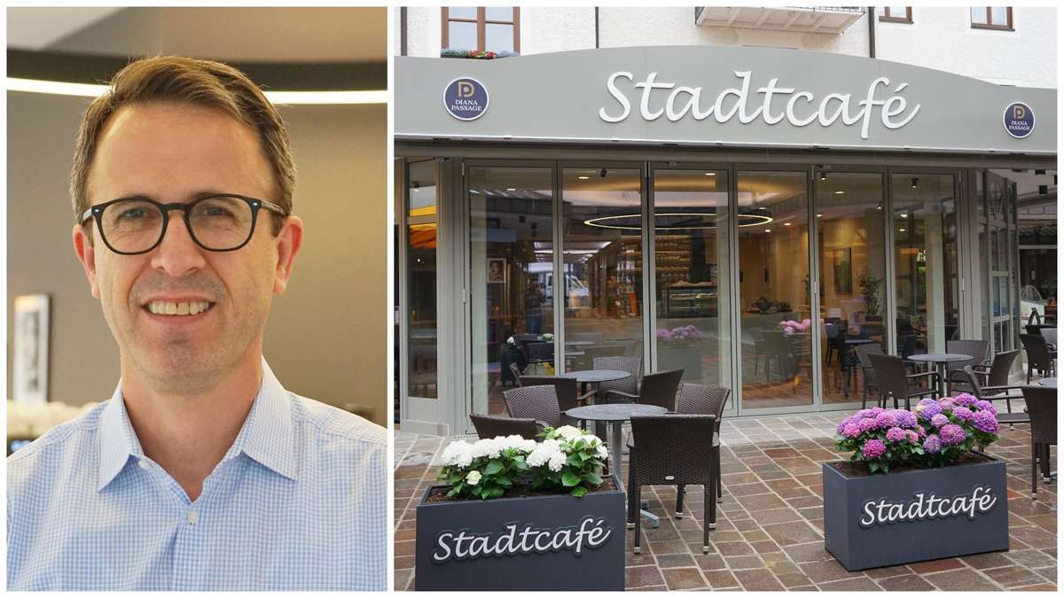 A man wearing glasses smiles beside an outdoor cafe. The cafe has glass doors, tables, and chairs. The cafe sign reads Stadtcafe.
