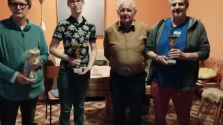Four men stand in a room holding trophies. They are dressed in casual clothes, with two older men wearing glasses. Behind them is a table with a lamp and a clock.
