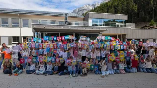 Eine große Gruppe von Kindern hält bunte Flaggen vor einem Gebäude mit Bergen im Hintergrund.