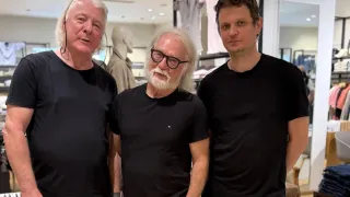 Drei Männer in schwarzen T-Shirts stehen in einem Bekleidungsgeschäft zusammen. Im Hintergrund steht ein Dummy in einem braunen Hemd.