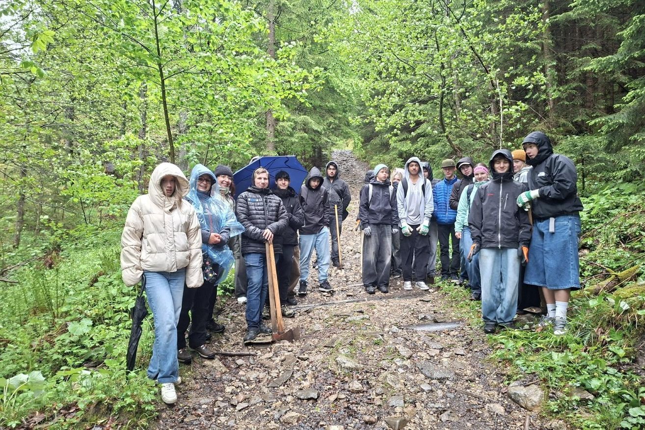 Eine Gruppe von Menschen in Regenkleidung steht auf einem Kiesweg im Wald, einige halten Werkzeuge. Sie scheinen an einem Gemeinschaftsprojekt oder Umweltprojekt zu arbeiten.