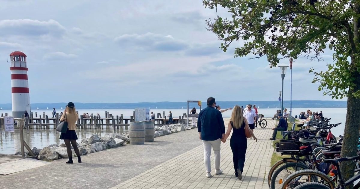 A couple walks hand-in-hand on a brick walkway by the lake. A woman in a skirt stands by the rocks, while others sit on benches. In the distance, there is a pier and boats on the water.