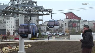 Eine Seilbahn ist von einem Kabel über einem Parkplatz mit vielen Autos aufgehängt. Eine Person steht in der Nähe der Seilbahn, und Gebäude sind im Hintergrund.