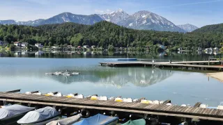 Ein ruhiger See mit einem Holzsteg, angedockten Booten und Bergen im Hintergrund. Bäume und Häuser befinden sich am Ufer.