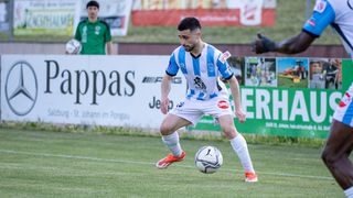 Fußballspieler im blau-weißen Trikot tritt den Ball. Im Hintergrund befinden sich Anzeigen für Pappas, ERHAU und andere.