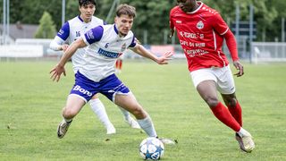 Drei Fußballspieler sind auf einem Rasenfeld. Zwei Spieler in weißen und blauen Uniformen jagen einem Spieler in einer roten Uniform nach. Der Spieler in der roten Uniform versucht, den Ball zu schießen.