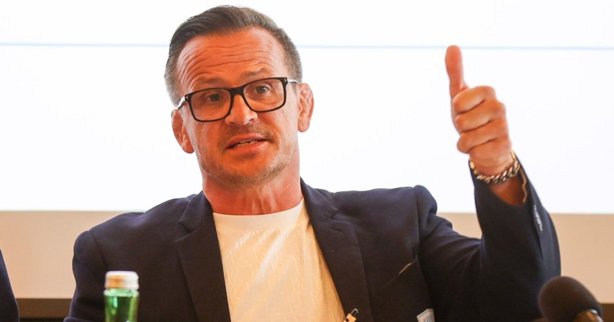 Ein Mann mit Brille und Anzug gibt einen Daumen hoch, mit einem verschwommenen Hintergrund.