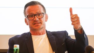 Ein Mann mit Brille und Anzug gibt einen Daumen hoch, mit einem verschwommenen Hintergrund.