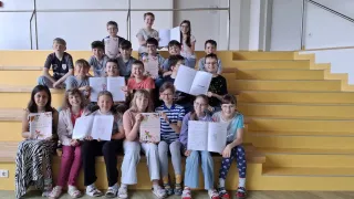 Eine Gruppe junger Schüler, Jungen und Mädchen, hält offene Bücher in den Händen und sitzt auf Tribünen, einige lächeln, andere posieren. Die Bücher scheinen Märchenbücher zu sein. Im Hintergrund befinden sich gelbe Tribünen und eine weiße Wand.