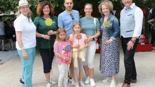 Eine Gruppe von Menschen steht lächelnd für ein Foto, einige halten Spielzeug. Die Frau links trägt eine Mütze und ein Armband. Die beiden Mädchen vorne haben Spielzeug. Dahinter sind Bäume und einige Fahnen.