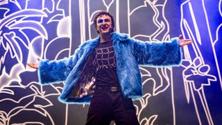 Ein lächelnder Mann in einem blauen Pelzmantel und schwarzem T-Shirt steht mit ausgestreckten Armen auf der Bühne. Er hat blaue Gesichtsbemalung.