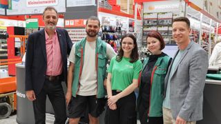 Fünf Personen in grünen Westen stehen in einem Geschäft. Drei lächeln, eine trägt eine Brille und ein Mann in einem rosa Hemd steht links.
