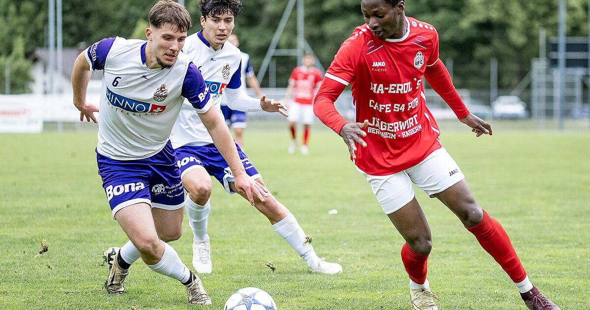 Drei Fußballspieler in verschiedenen Trikots laufen auf einem Grasfeld. Einer ist in Rot, ein anderer in Weiß und Blau und der dritte in Weiß. Ein Spieler ist kurz davor, den Ball zu treten.