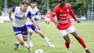 Drei Fußballspieler in verschiedenen Trikots laufen auf einem Grasfeld. Einer ist in Rot, ein anderer in Weiß und Blau und der dritte in Weiß. Ein Spieler ist kurz davor, den Ball zu treten.