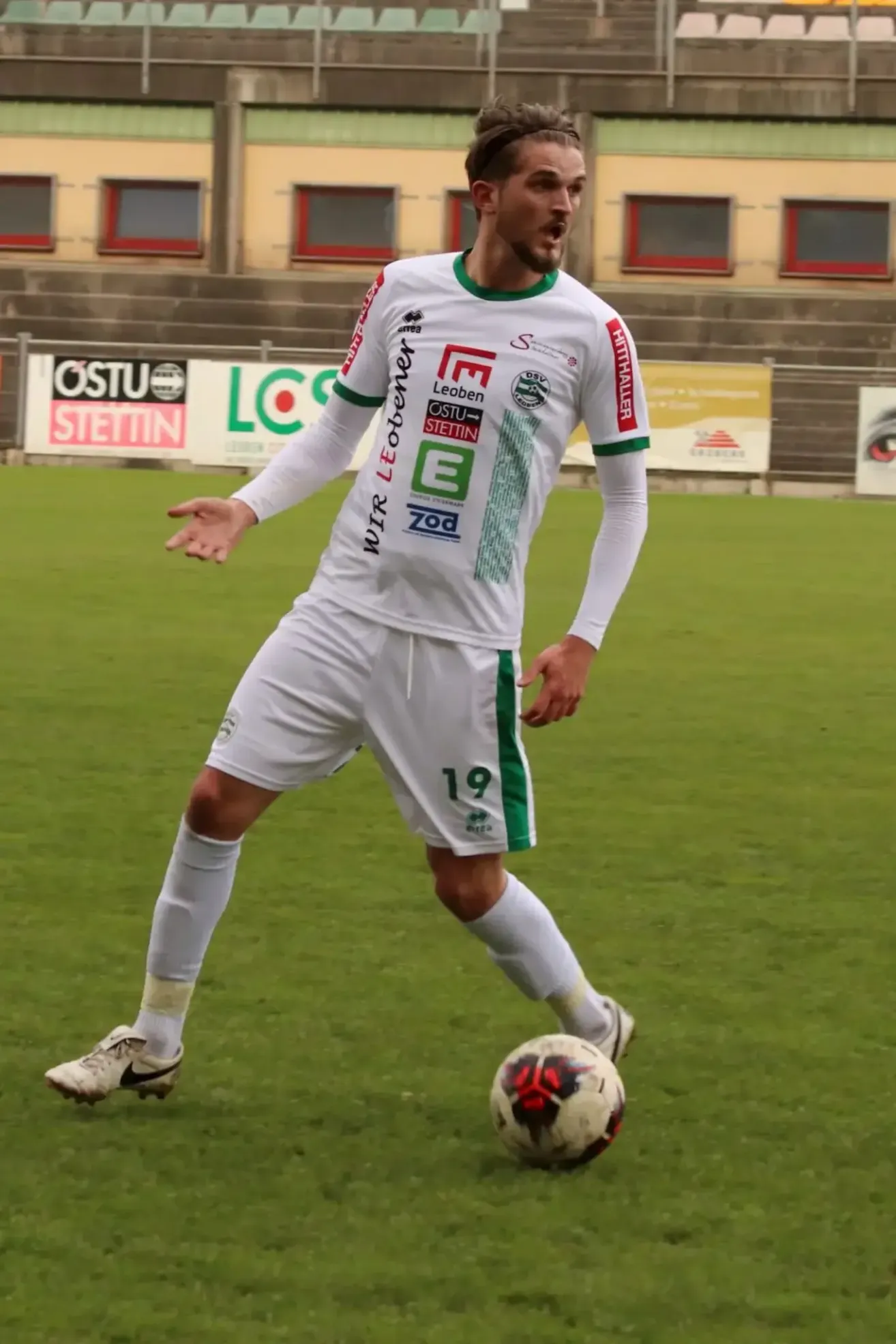 Ein Fußballspieler in einem weißen und grünen Trikot steht auf einem Feld, bereit einen Fußball zu treten.