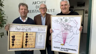 Drei Männer stehen im Innenbereich und lächeln. Sie halten gerahmte Dokumente und eine Karte. Der linke Mann hält ein Dokument mit dem Titel 'Staatsvertrag 1955'. Der mittlere Mann hält eine Karte mit dem Titel 'Ruster Wein-Feuerhardel'. Der rechte Mann hält ein Dokument und eine Karte. Hinter ihnen steht eine Wand mit der Aufschrift 'Archiv Di Reistal Rust.'