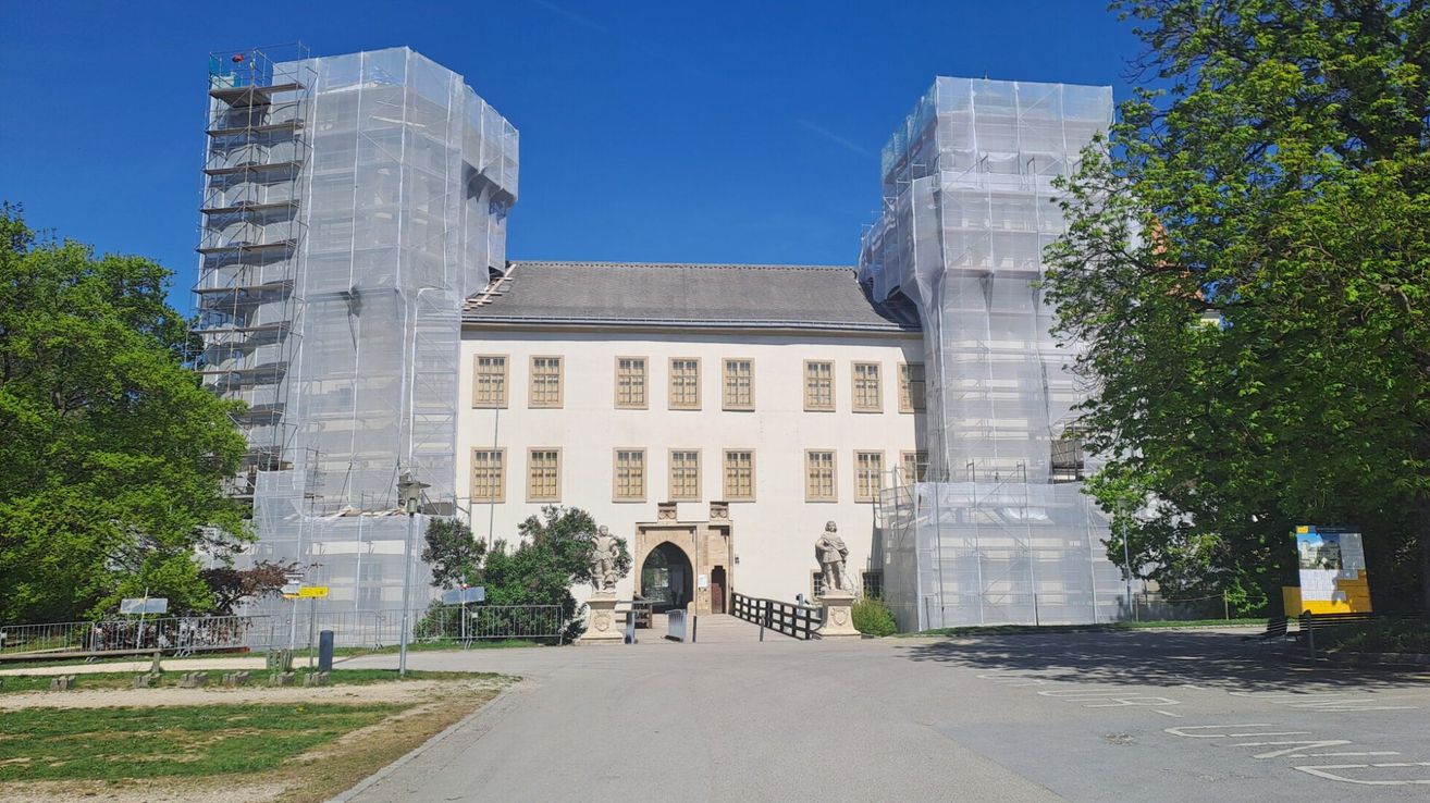 Ein weißes Gebäude mit grauem Dach und vielen Fenstern wird renoviert. Zwei Statuen und ein Zaun stehen vor dem Eingang.