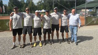 Sechs Männer in identischen weißen T-Shirts und schwarzen Shorts posieren für ein Foto auf einer Kiesfläche mit einem Tennisplatz im Hintergrund.