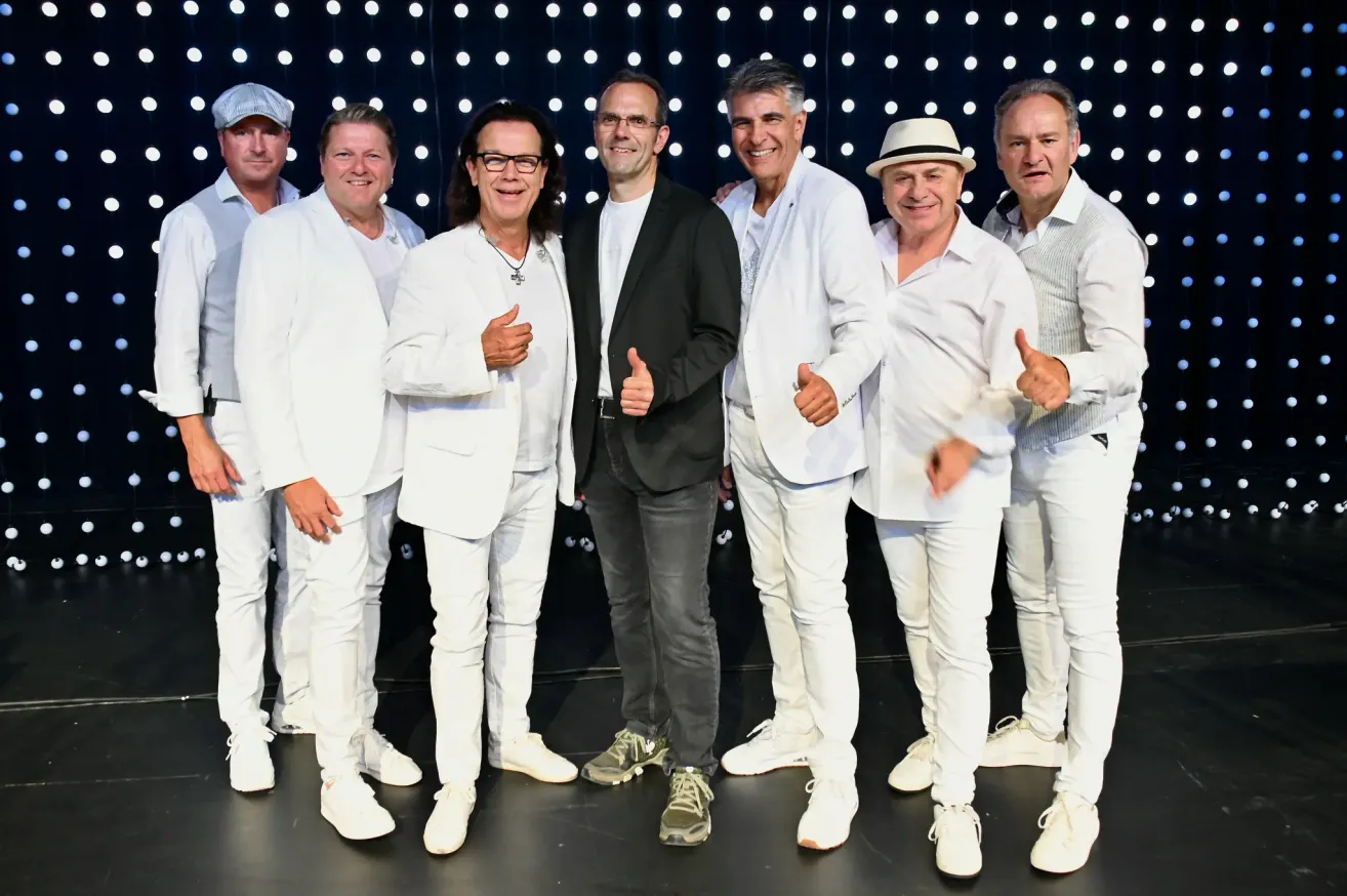 Eine Gruppe von Männern in weißen Anzügen, Hüten und Sneakers lächelt und posiert für ein Foto auf einer Bühne.