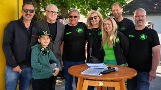 Eine Gruppe lächelnder Erwachsener und eines Kindes steht um einen Tisch mit Mikrofonen und Papieren. Sie tragen passende grüne Shirts. Bäume und ein Gebäude sind im Hintergrund.