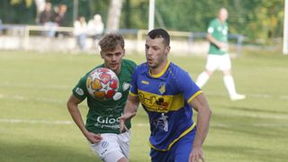 Zwei Fußballspieler auf einem Feld, einer in Grün und einer in Blau. Der Spieler in Grün ist dabei, einen bunten Fußball zu treten. Der Spieler in Blau versucht, ihn zu blocken.