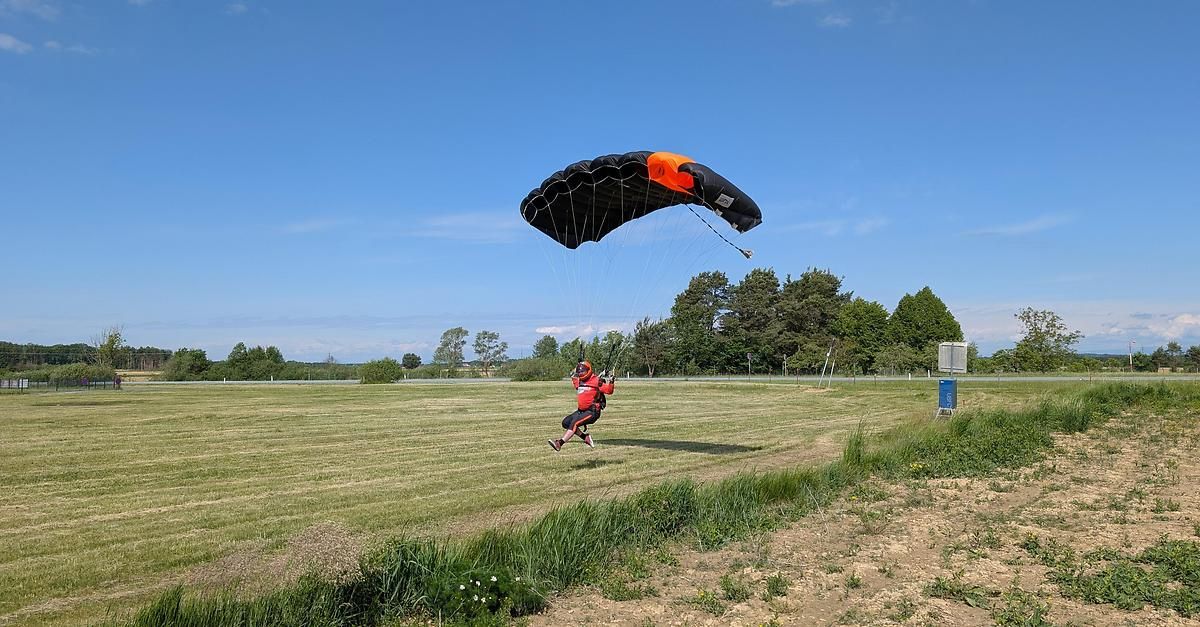 Eine Person mit einem roten Outfit und Helm fällt mit einem schwarzen und orangefarbenen Fallschirm über ein grasiges Feld.