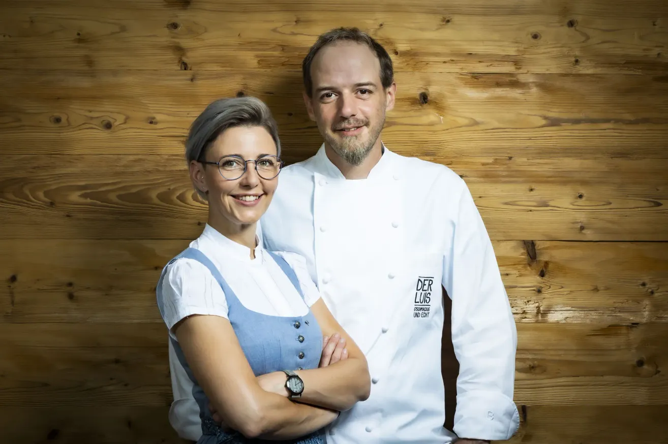Ein Mann und eine Frau, beide in weißen Kochuniformen, stehen lächelnd mit verschränkten Armen vor einer Holzwand.