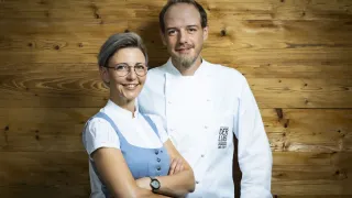 Ein Mann und eine Frau, beide in weißen Kochuniformen, stehen lächelnd mit verschränkten Armen vor einer Holzwand.