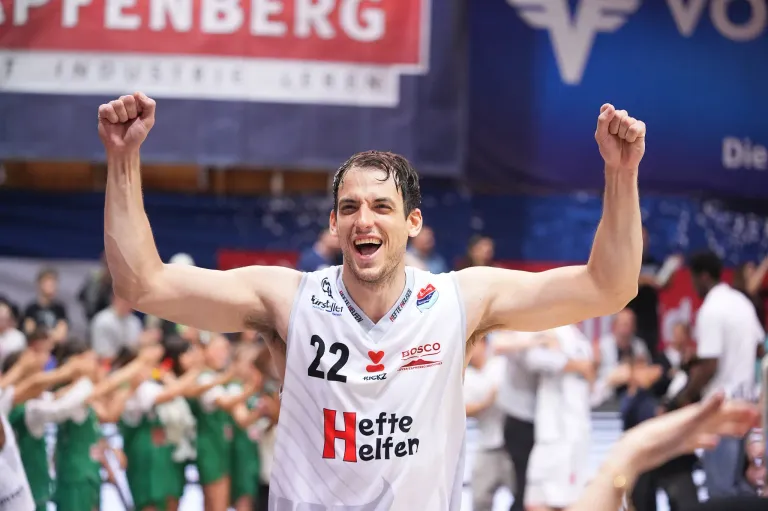 Ein Basketballspieler feiert den Sieg vor einer Menschenmenge. Er trägt ein weißes Trikot mit der Nummer 22 und dem Wort Heffe Helfen.