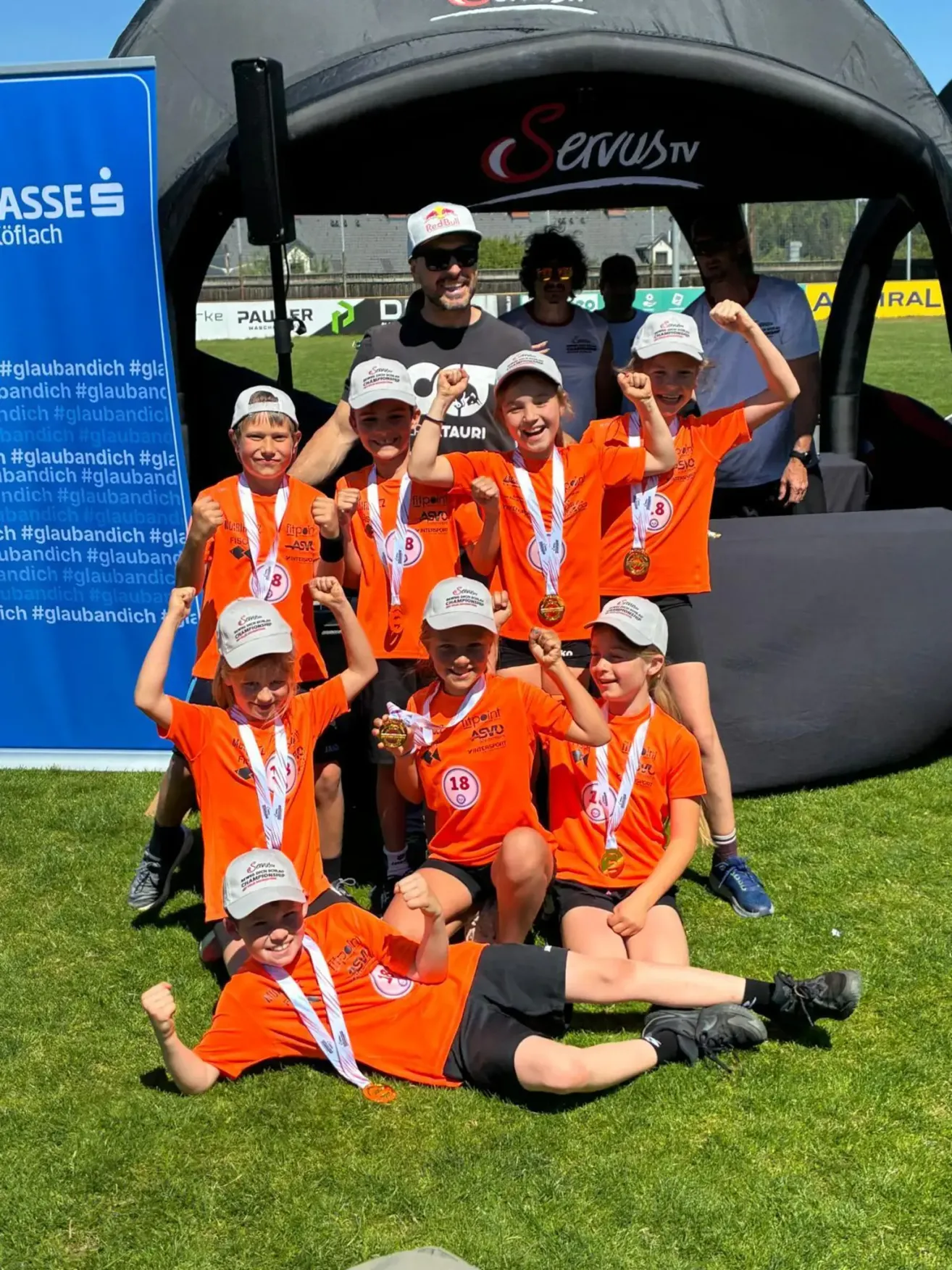 Gruppe von Kindern in orangefarbenen Uniformen mit Medaillen feiert auf einem Feld mit einem Zelt und Erwachsenen im Hintergrund.