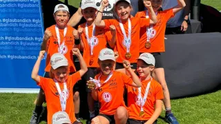 Gruppe von Kindern in orangefarbenen Uniformen mit Medaillen feiert auf einem Feld mit einem Zelt und Erwachsenen im Hintergrund.