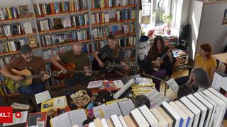 Vier Personen spielen Gitarren in einem von Büchern umgebenen Raum. Einer spielt Ukulele, und sie alle lächeln. Es gibt Bücher auf dem Tisch und in den Regalen hinter ihnen.
