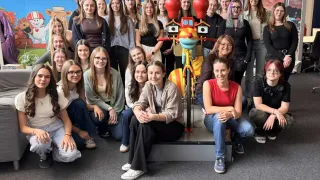 Eine Gruppe von Frauen posiert für ein Foto, einige sitzend und andere stehend, um eine große roboterähnliche Figur in einer Studioumgebung mit einem Hintergrund, der 'okiDoki' und Luftballons zeigt.