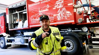 Ein Mann in einer reflektierenden Weste hält ein Funkgerät vor einem Feuerwehrauto.