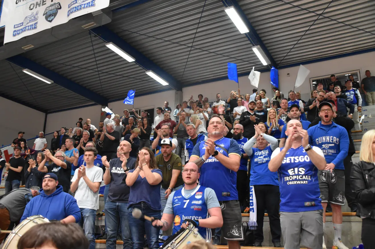 Eine Menschenmenge in blauen Trikots jubelt während einer Sportveranstaltung. Einige halten blaue Fahnen und klatschen begeistert. Eine Person in der Front spielt eine Trommel und steigert die Aufregung.