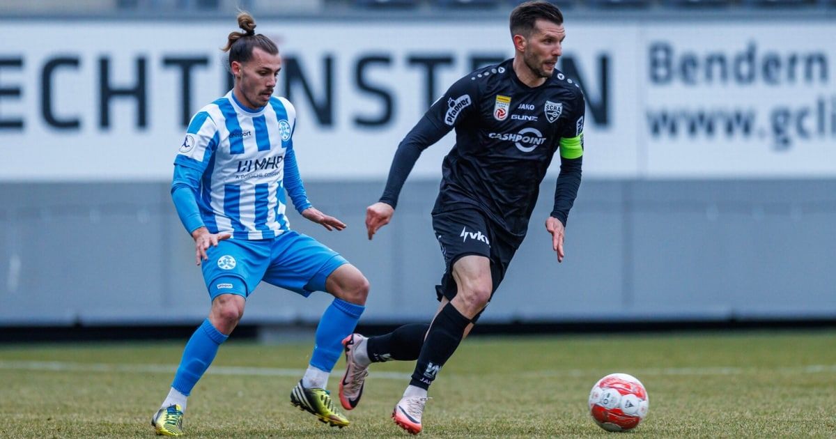 Zwei Fußballspieler konkurrieren auf dem Feld. Einer trägt ein blau-weißes Trikot, der andere ein schwarzes. Der Spieler im schwarzen Trikot jagt dem Ball nach.