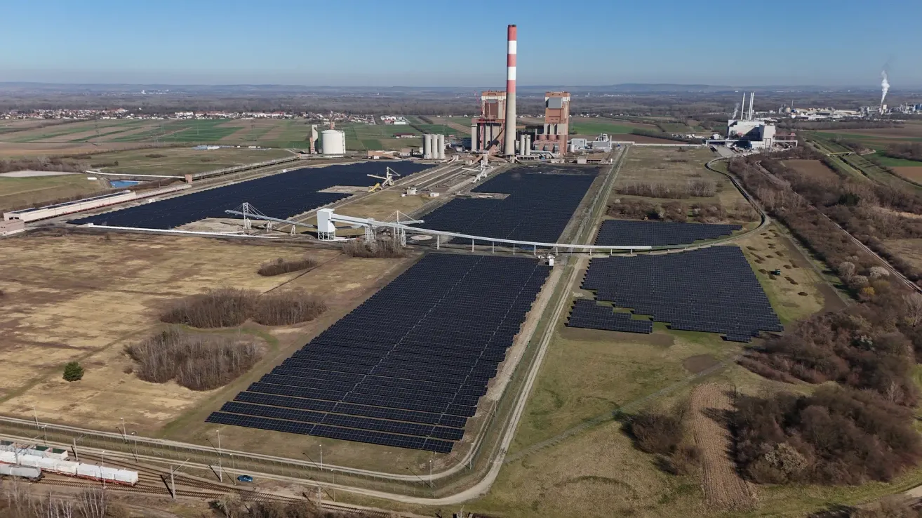 Luftaufnahme eines großen Solarkraftwerks mit Solarmodulen und einem Schornstein. Im Hintergrund sind Industriegebäude und grüne Felder zu sehen.