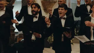 Eine Gruppe formell gekleideter Männer singt in einer Kirche mit einem Kreuz und Blumen im Hintergrund.