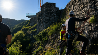 Drei Personen klettern an einer steilen Wand und tragen Helme und Gurte. Eine Person steht am Boden und beobachtet.