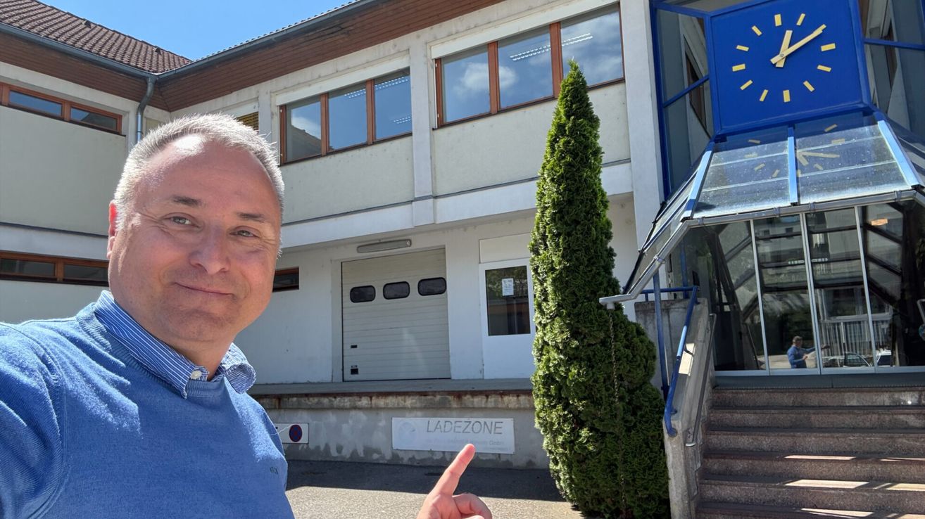Ein Mann steht lächelnd vor einem Gebäude mit weißer Außenwand und blauer Einfassung und zeigt mit dem Finger. Das Gebäude hat eine Garagentür, Fenster und ein Schild mit der Aufschrift 'LADEZONE'. Rechts steht ein großer grüner Busch.