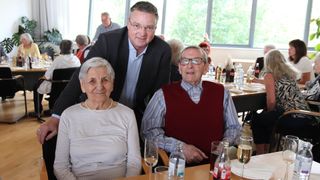 Ein älteres Paar, beide mit Brille, sitzt an einem Tisch. Hinter ihnen steht ein weiterer Mann. Auf dem Tisch befinden sich Flaschen, Gläser und Teller, und andere Menschen sitzen im Hintergrund.