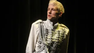 Ein Mann mit blonden Haaren steht auf der Bühne und trägt eine weiße und goldene Uniform mit einem Mikrofon am Ohr.