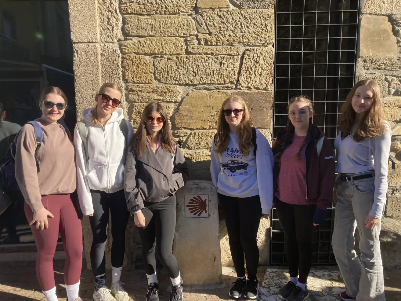 Fünf junge Frauen stehen vor einer Steinmauer mit einem roten Schild. Sie tragen alle Sonnenbrillen und Turnschuhe, und eine trägt eine Jacke. Die Mauer hat ein Metallgitter.
