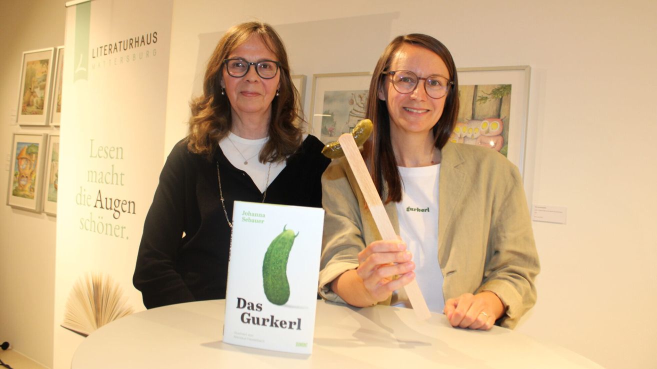 Zwei Frauen sitzen an einem Tisch mit einem Buch namens 'Das Gurkerl' und einem Gurken. Sie lächeln und posieren für ein Foto.