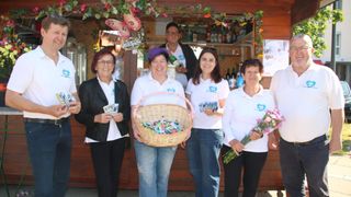 Fünf Frauen stehen lächelnd vor einem hölzernen Tresen. Eine hält einen Korb mit Süßigkeiten, eine andere Blumen. Hinter ihnen hält ein Mann eine Flasche. Mit Flaschen und Gegenständen bestückte Regale schmücken den Tresen.