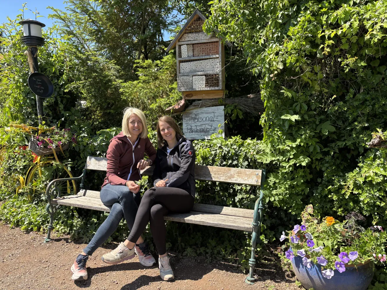 Zwei Frauen sitzen auf einer Bank in einem Garten mit einem Schild, das 'Personal Parking' steht. Sie lächeln und eine hält eine Pflanze. Ein Bienenstock hängt über ihnen, umgeben von üppiger Grünfläche.