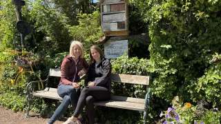 Zwei Frauen sitzen auf einer Bank in einem Garten mit einem Schild, das 'Personal Parking' steht. Sie lächeln und eine hält eine Pflanze. Ein Bienenstock hängt über ihnen, umgeben von üppiger Grünfläche.