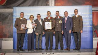 Eine Gruppe von Menschen steht mit Auszeichnungen und einem Zertifikat auf der Bühne, trägt Militäruniformen und formelle Kleidung. Ein Mann hält ein Zertifikat, und eine Frau hält eine Auszeichnung.