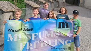 Fünf Kinder halten ein Banner vor einem Museum. Der Spruch auf dem Banner 'Museumsfruhling' ist in weißen Buchstaben auf einem bunten Hintergrund. Die Kinder lächeln und scheinen für ein Foto zu posieren.