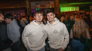 Zwei junge Männer mit Brillen und Kapuzenpullovern stehen lächelnd nebeneinander bei einer überfüllten Veranstaltung. Hinter ihnen tummeln sich eine Menge Menschen, und ein Schild sagt 'Maiwiperl Party'.